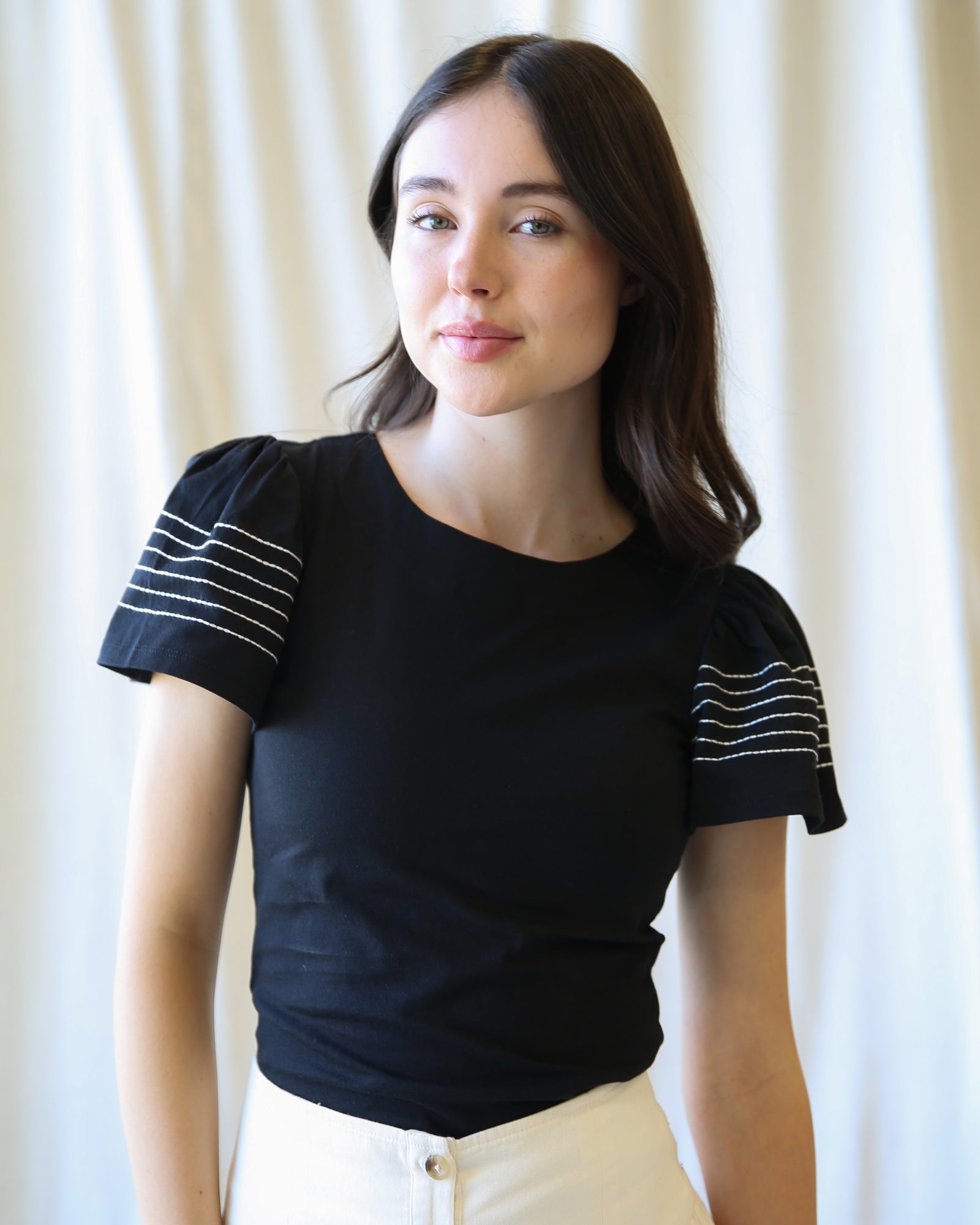 Woman wearing a black top with white stripes on the sleeves against a white curtain background