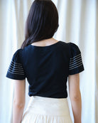 Person wearing a black top with white stripes on sleeves against a white curtain background