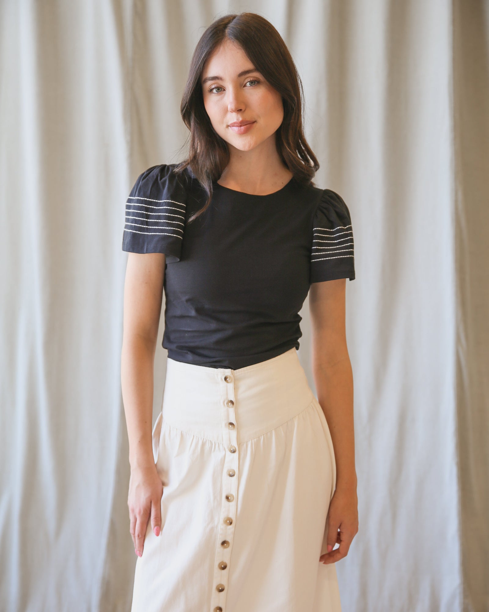 Woman wearing a black top with striped sleeves and a beige skirt against a plain background