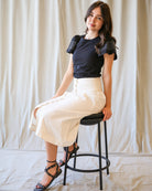 Woman sitting on a stool wearing a black top and white pants against a plain background