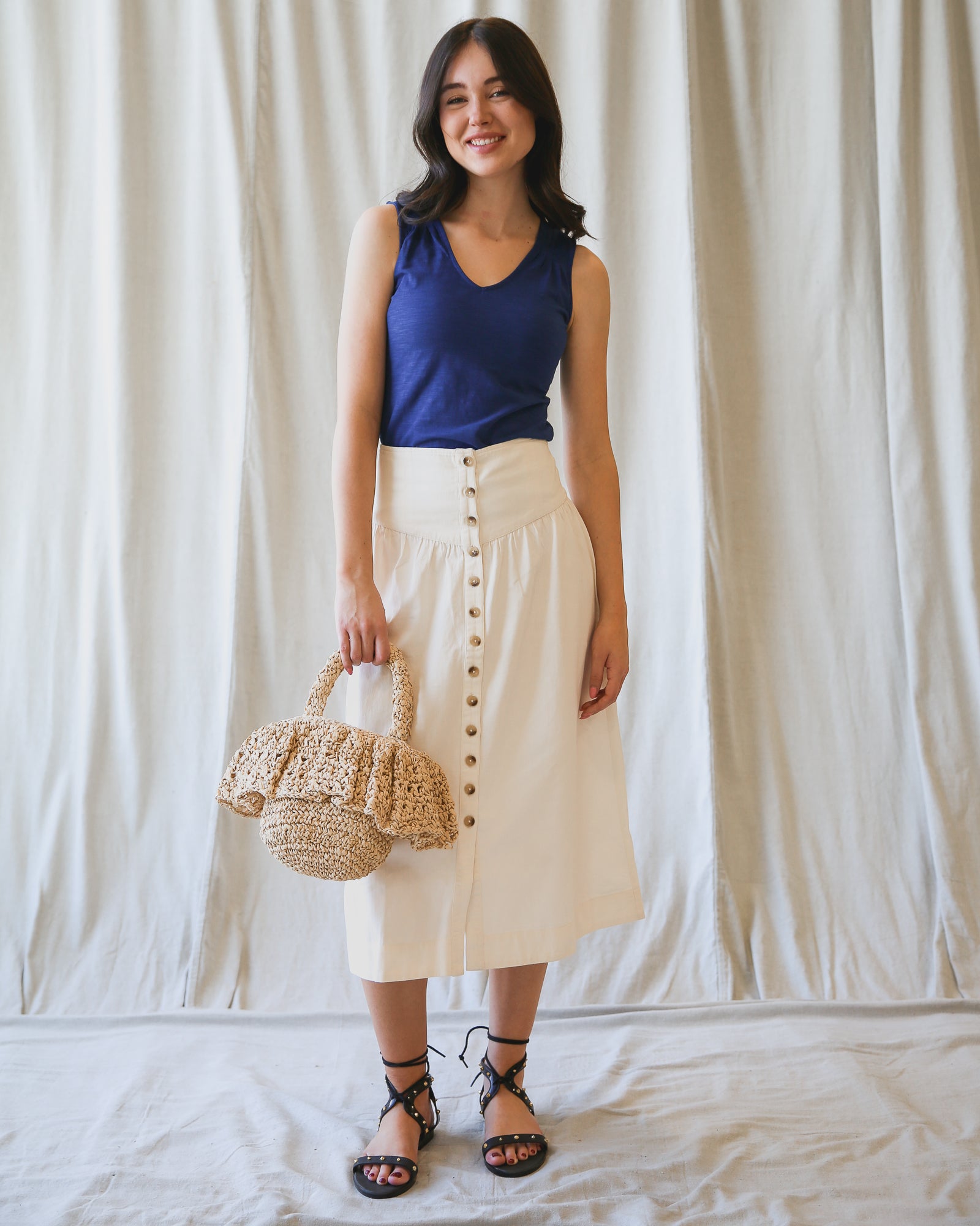 Woman wearing a blue top and white skirt holding a straw bag against a plain background