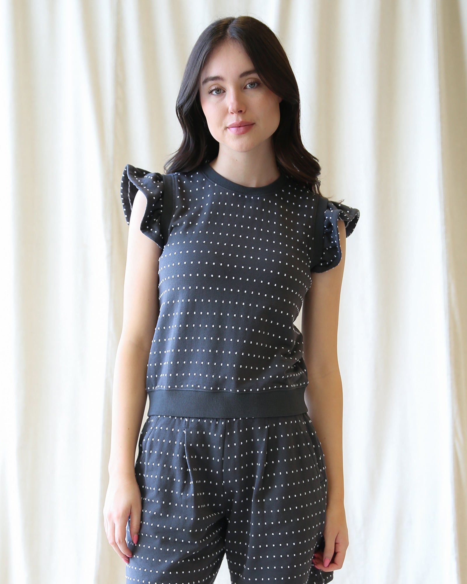 Woman wearing a dark blue top with white polka dots and matching pants against a white curtain background