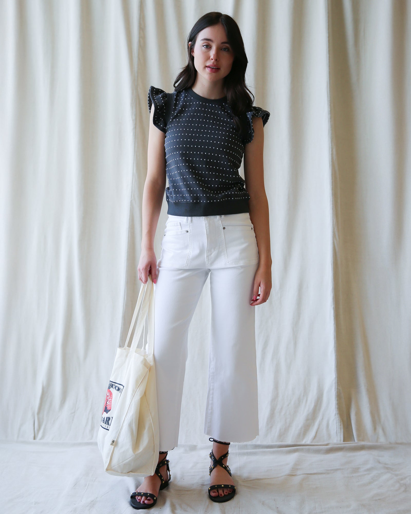 Woman wearing a black patterned top and white pants, holding a white tote bag against a plain background