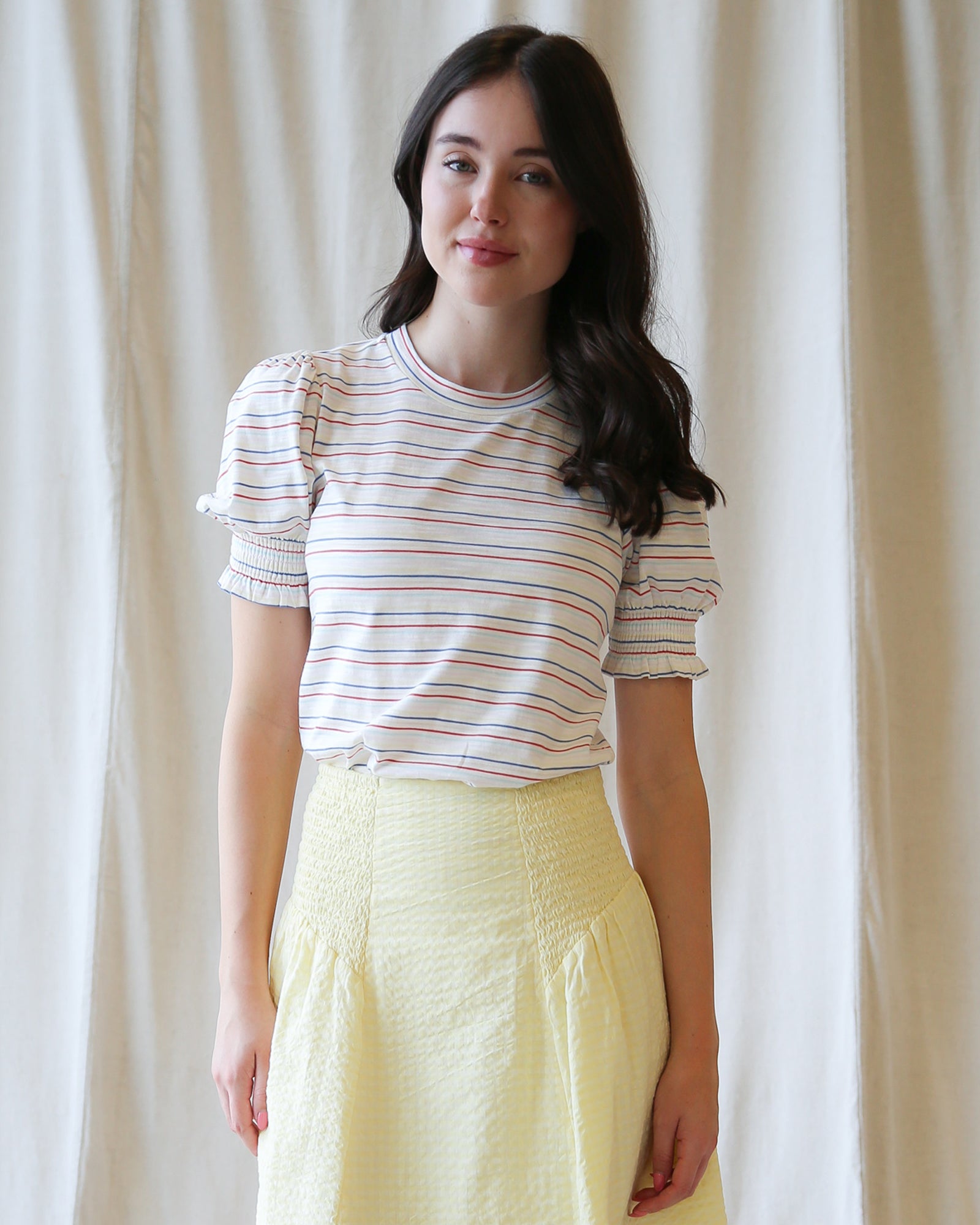 Woman wearing a striped shirt and yellow pants against a white curtain background