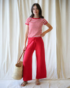 Woman wearing a red and white striped shirt and red pants, holding a straw bag against a white curtain background.