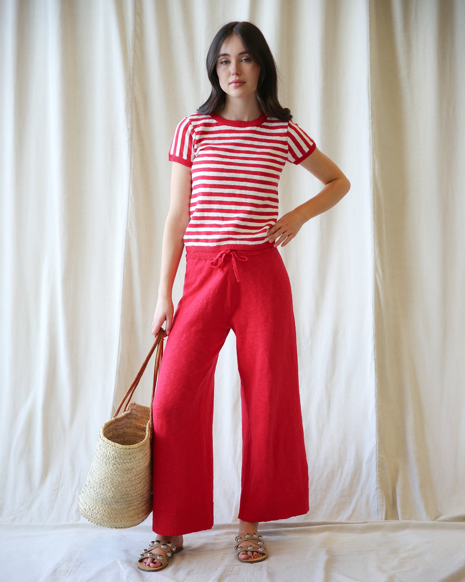 Woman wearing a red and white striped shirt and red pants, holding a straw bag against a white curtain background.