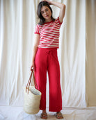 Woman wearing a red and white striped shirt and red pants, holding a straw bag against a white curtain background.