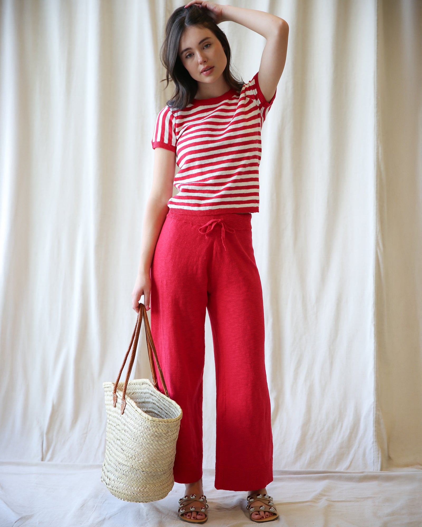 Woman wearing a red and white striped shirt and red pants, holding a straw bag against a white curtain background.