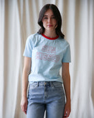 Person wearing a light blue t-shirt with 'SUN KISSED' text and jeans against a white curtain background