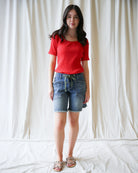 Woman wearing a red top and denim shorts against a white curtain background
