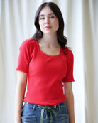 Woman wearing a red top and blue jeans against a white curtain background