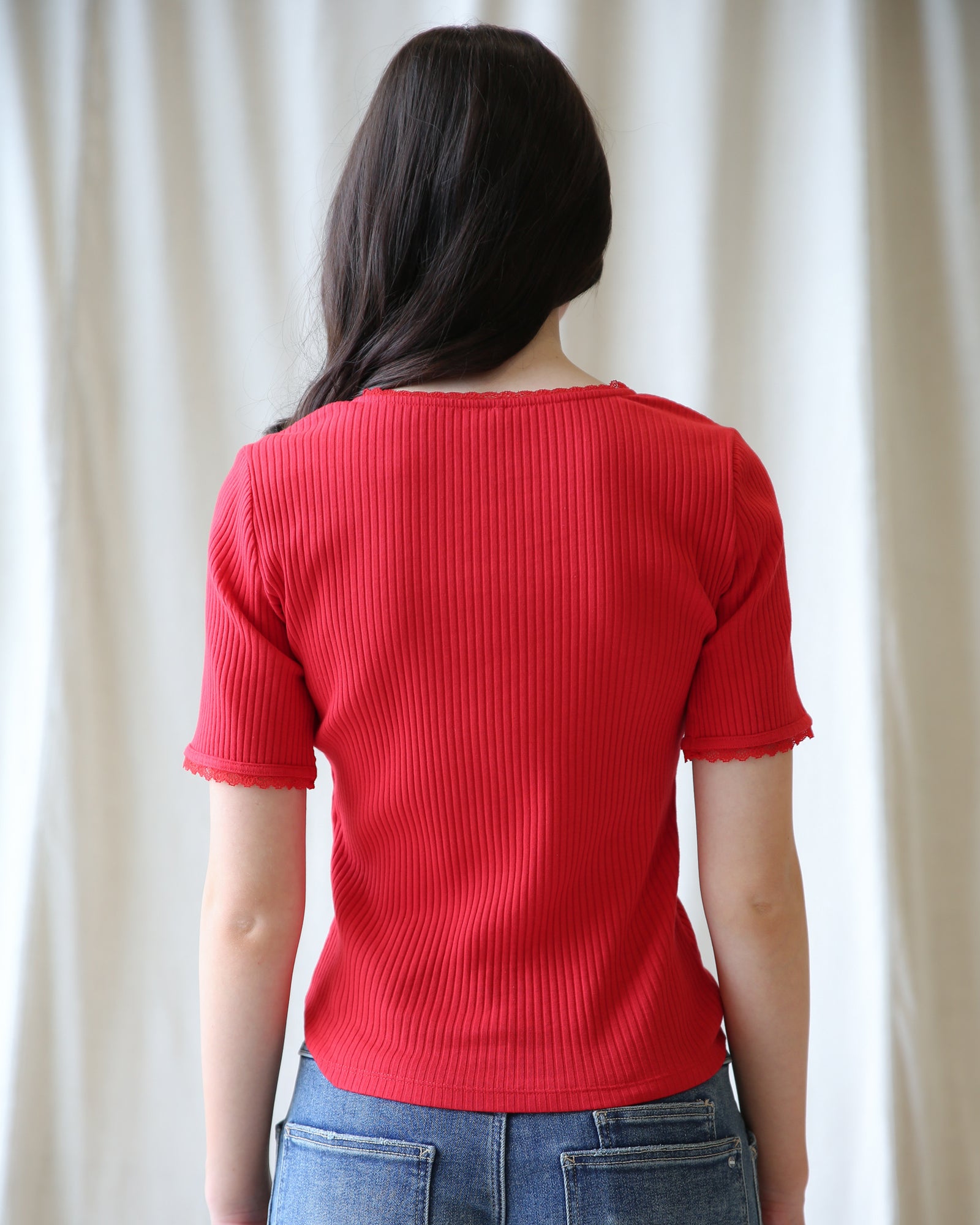 Person wearing a red ribbed sweater and blue jeans against a white curtain background