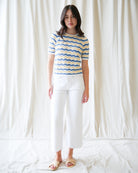 Woman wearing a blue and yellow striped shirt and white pants against a white curtain background