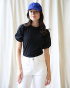 Woman wearing a black top and white pants with a blue cap against a white curtain background