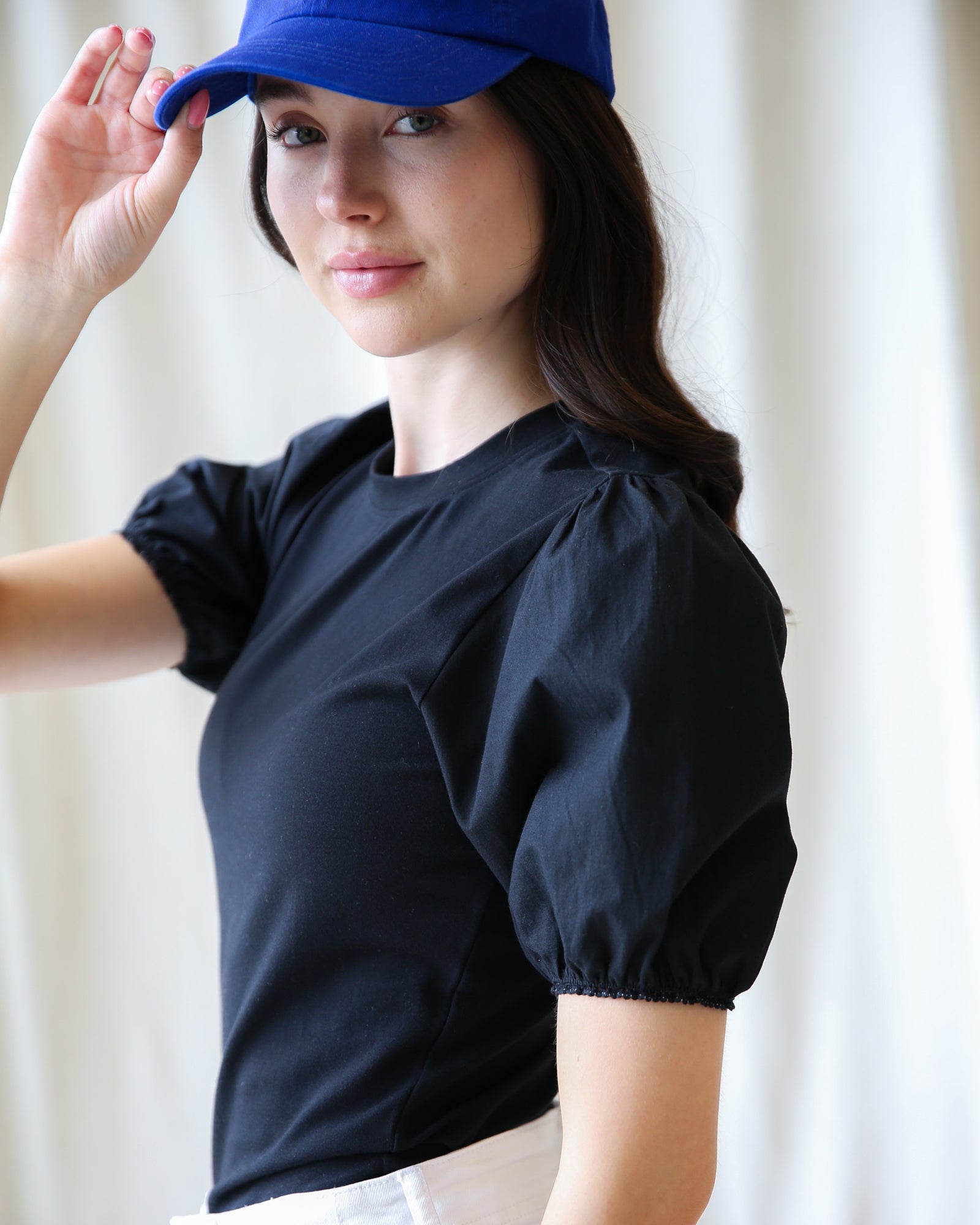 Woman wearing a black top with puff sleeves and a blue cap against a white curtain background