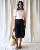 Woman wearing a white top with pink sleeves and a black skirt, holding a woven bag against a plain background.