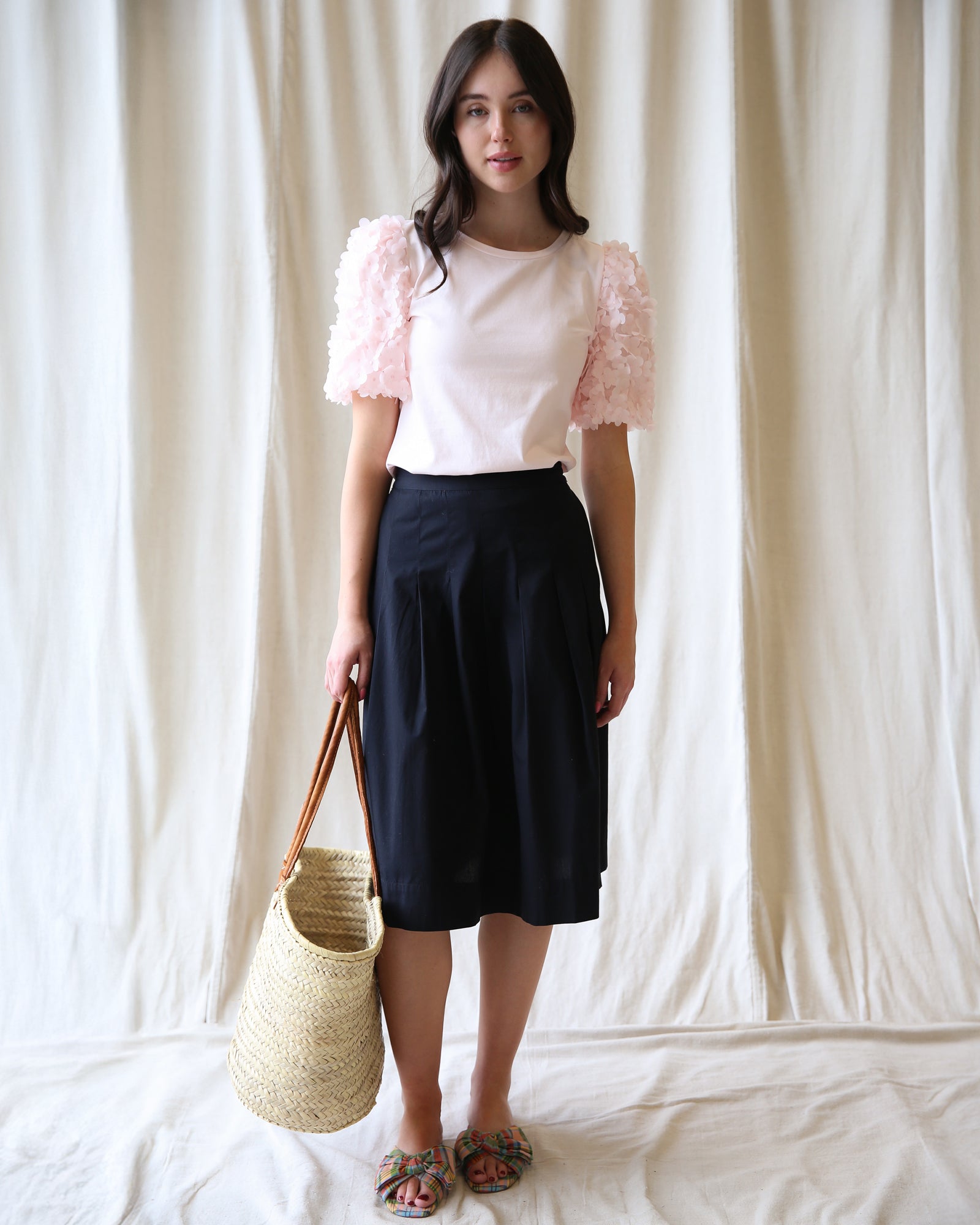 Woman wearing a white top with pink sleeves and a black skirt, holding a woven bag against a plain background.