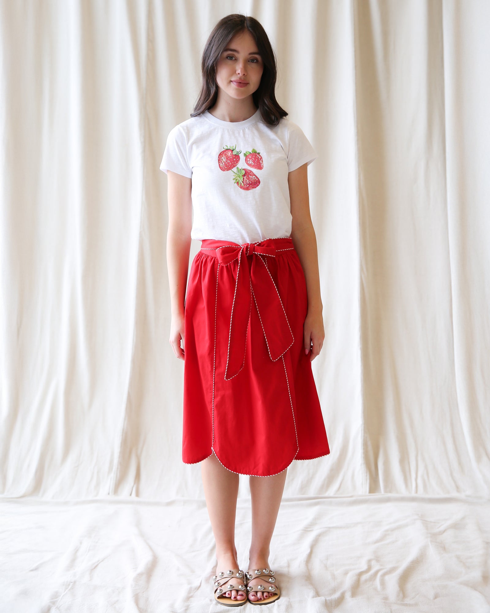 Woman wearing a white t-shirt with strawberry design and a red skirt against a plain background