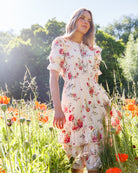 A woman stands in a sunlit field of orange flowers wearing a cream-colored, short-sleeved tiered dress with a red and pink floral print.