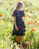 A young girl wearing a dark navy blue floral dress stands in a sunny grassy field filled with orange poppies.