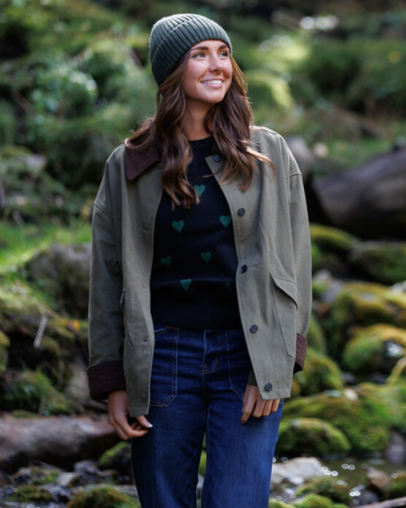 Woman standing in a forest wearing a green jacket, black sweater with heart patterns, and blue jeans.