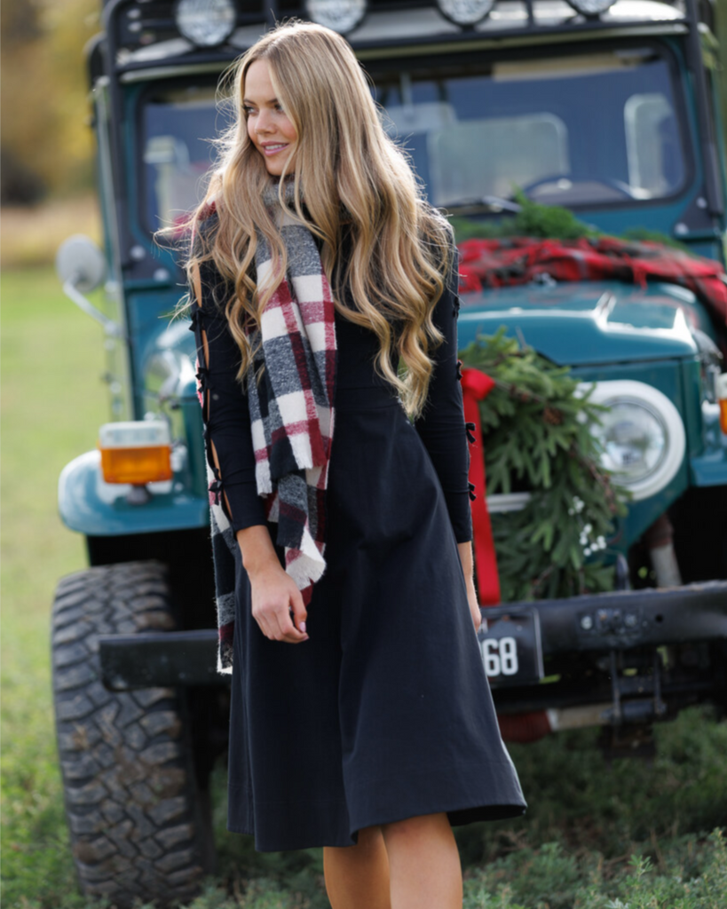 Woman wearing a black dress and plaid scarf standing in front of a teal SUV with Christmas decorations.