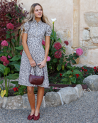 Woman in a patterned dress standing in a garden with flowers and stone architecture.