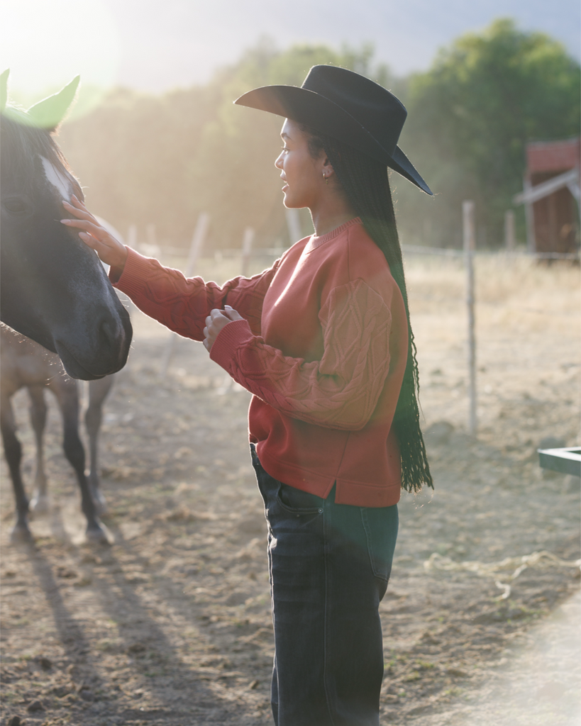 Woman in a cowboy hat and orange sweater interacting with a horse in a rural setting