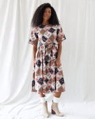 A woman modeling a short-sleeve midi dress with a multicolor patchwork diamond pattern in beige, navy, red, and white, styled with white socks and beige ankle boots against a white draped fabric background.