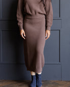 A person wearing a ribbed knit two-piece sweater and midi skirt set in a dusty brown color, paired with navy blue ankle boots, standing against a dark blue paneled wall.