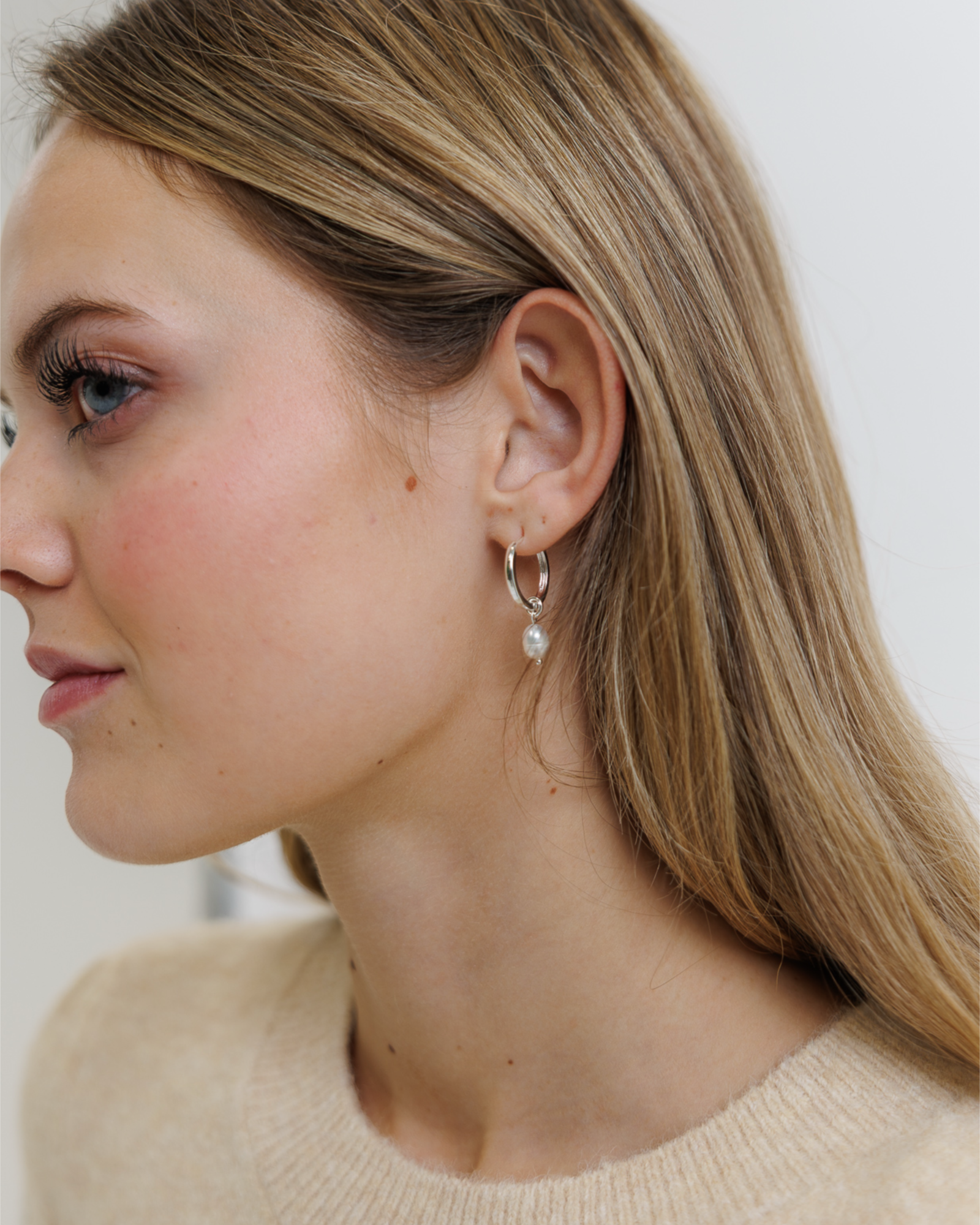 Close-up of a woman wearing silver hoop earrings with a pearl charm on a neutral background