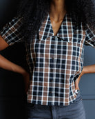 A woman wearing a short-sleeve plaid button-up shirt with a collar, featuring a pattern of brown, navy, white, and black. She is also wearing dark gray jeans, standing against a dark gray wall.