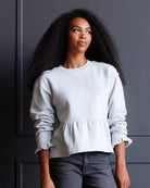 A woman with long, curly black hair wearing a light blue-gray crewneck sweatshirt with ruffled cuffs and a peplum hem, paired with dark gray jeans, standing against a dark gray paneled wall.