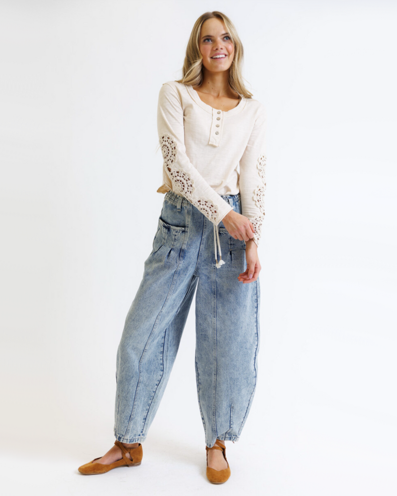 Woman wearing a cream colored long sleeve with crochet detailed sleeves and light denim pants against a white background