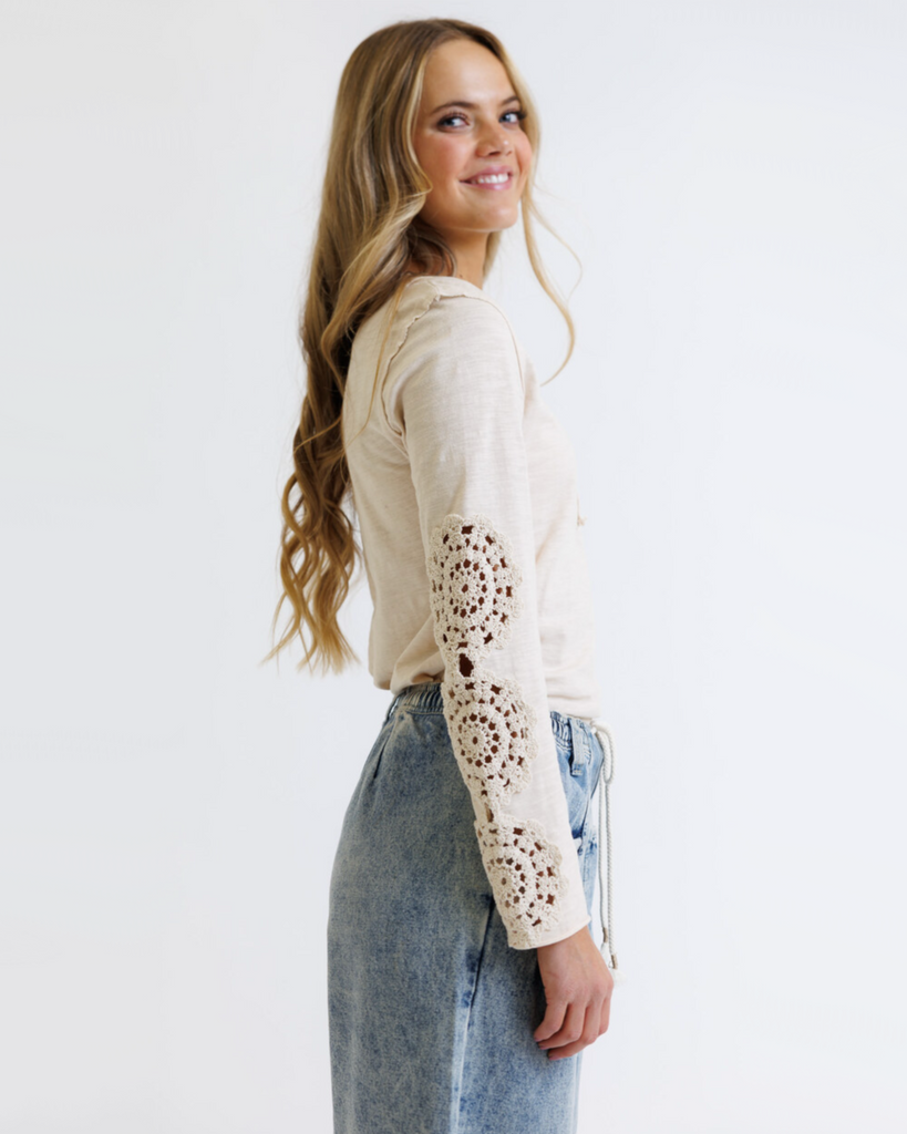 Woman wearing a cream colored long sleeve with crochet detailed sleeves against a white background