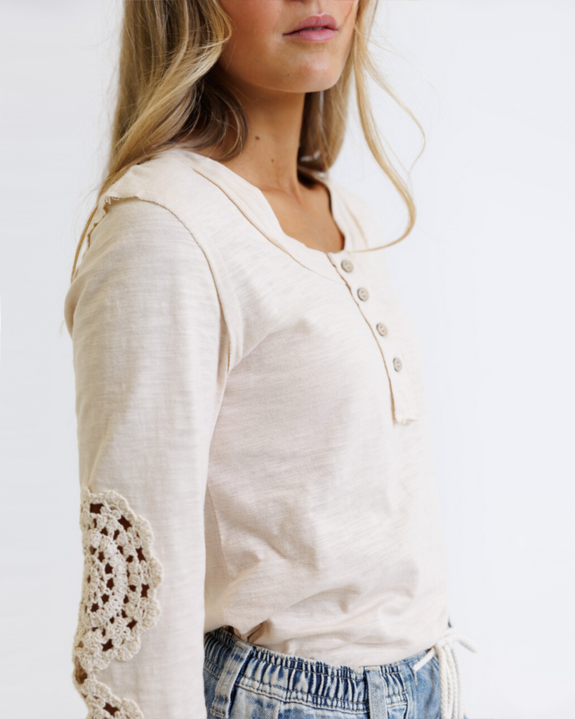 Woman wearing a cream colored long sleeve with crochet detailed sleeves against a white background