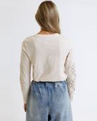 Woman wearing a cream colored long sleeve with crochet detailed sleeves against a white background
