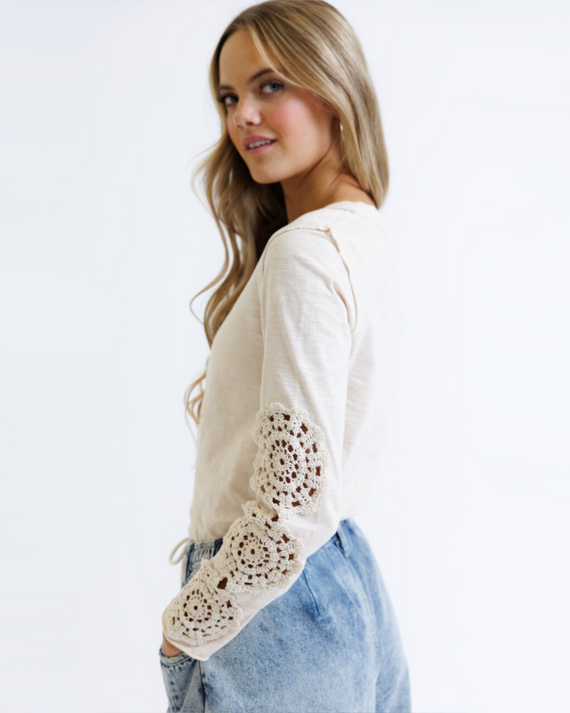 Woman wearing a cream colored long sleeve with crochet detailed sleeves against a white background