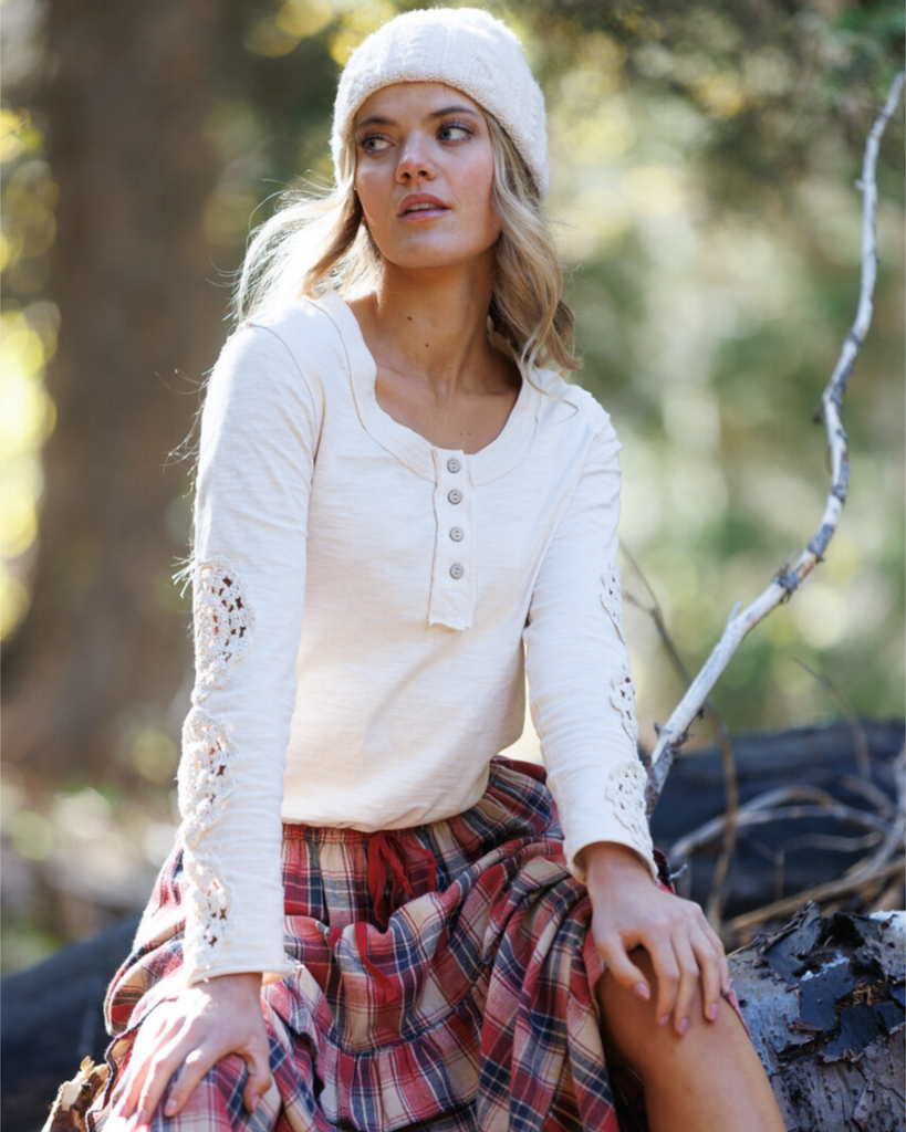 Woman sitting in a forest wearing a cream colored long sleeve and red plaid skirt