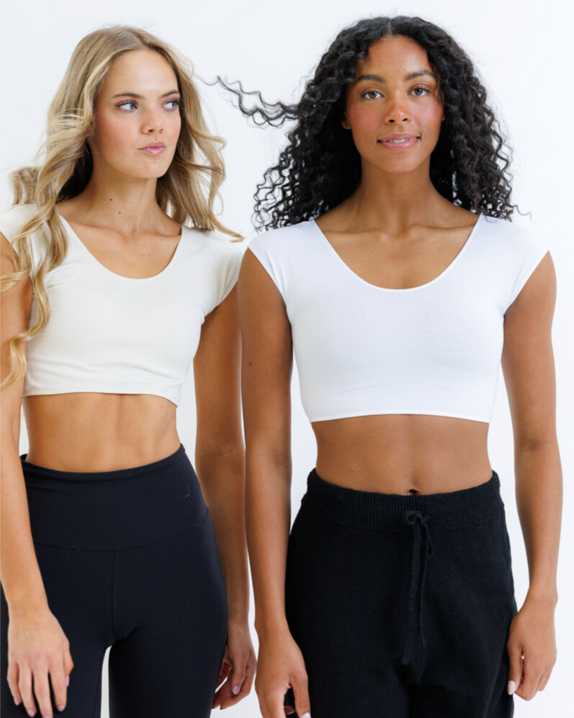 Two women wearing white cropped layering tops and black leggings on a plain background