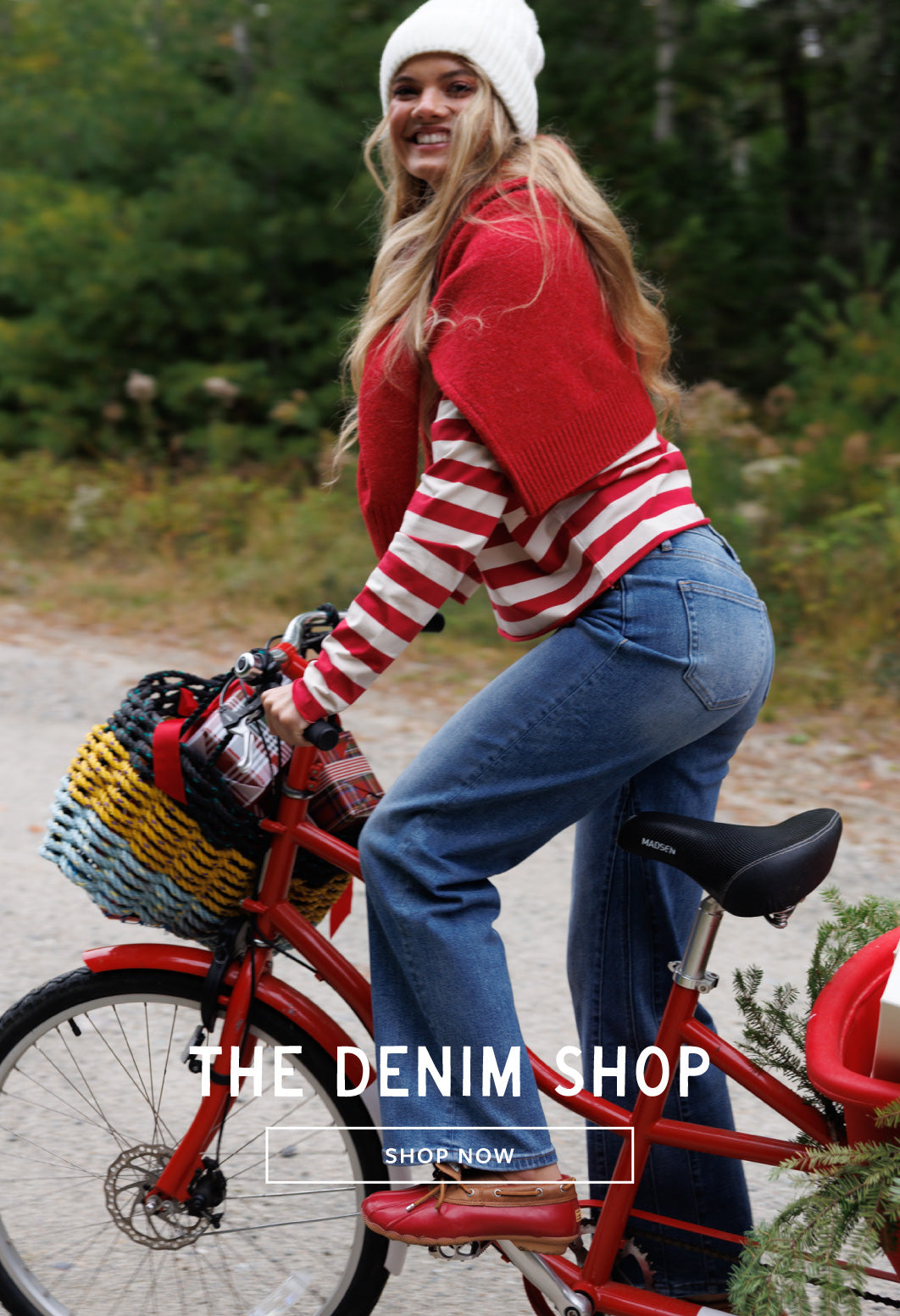 A woman riding a red bicycle outdoors, wearing a red and white striped sweater, blue jeans, and a white beanie. The text 'THE DENIM SHOP' is overlaid at the bottom.