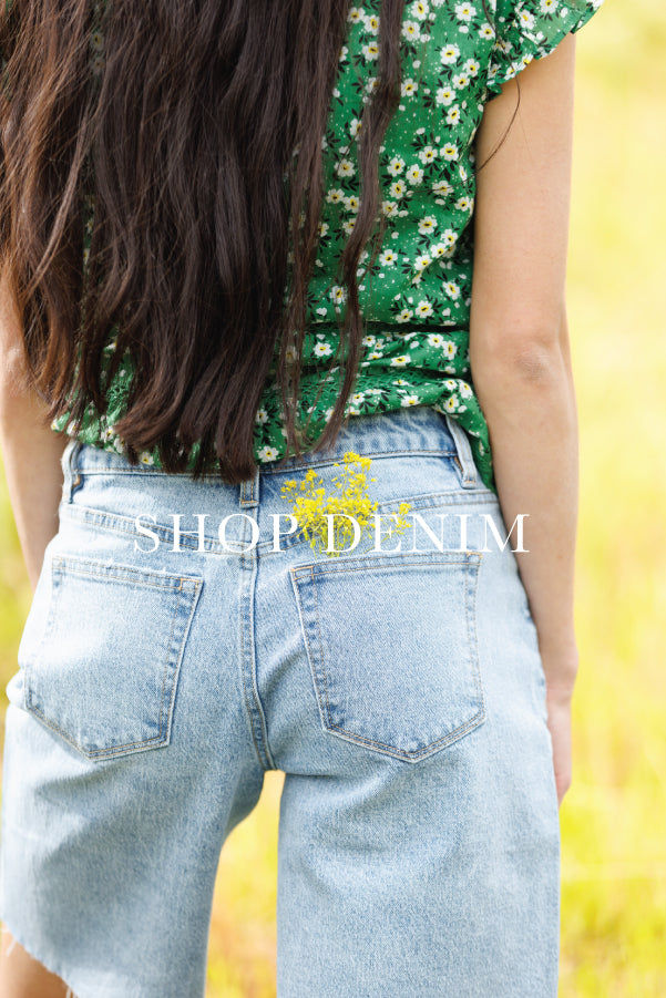 Rear view of a person wearing light-wash denim shorts and a green floral top, with a large 'SHOP DENIM' watermark across the center.