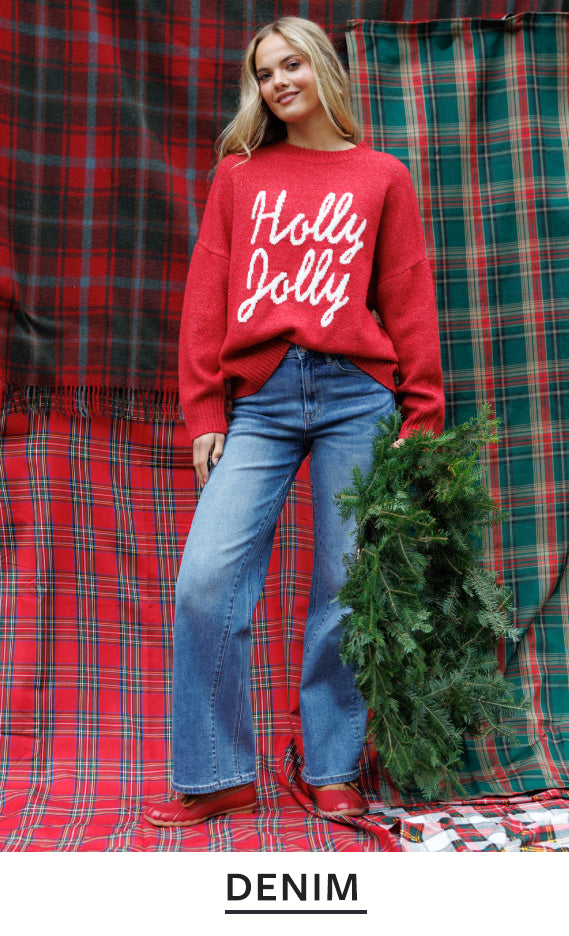 A woman stands in front of a red and green plaid backdrop wearing a red crewneck sweatshirt with 'Holly Jolly' in white script, blue jeans, and red shoes, while holding a green pine branch. The word 'DENIM' appears at the bottom.
