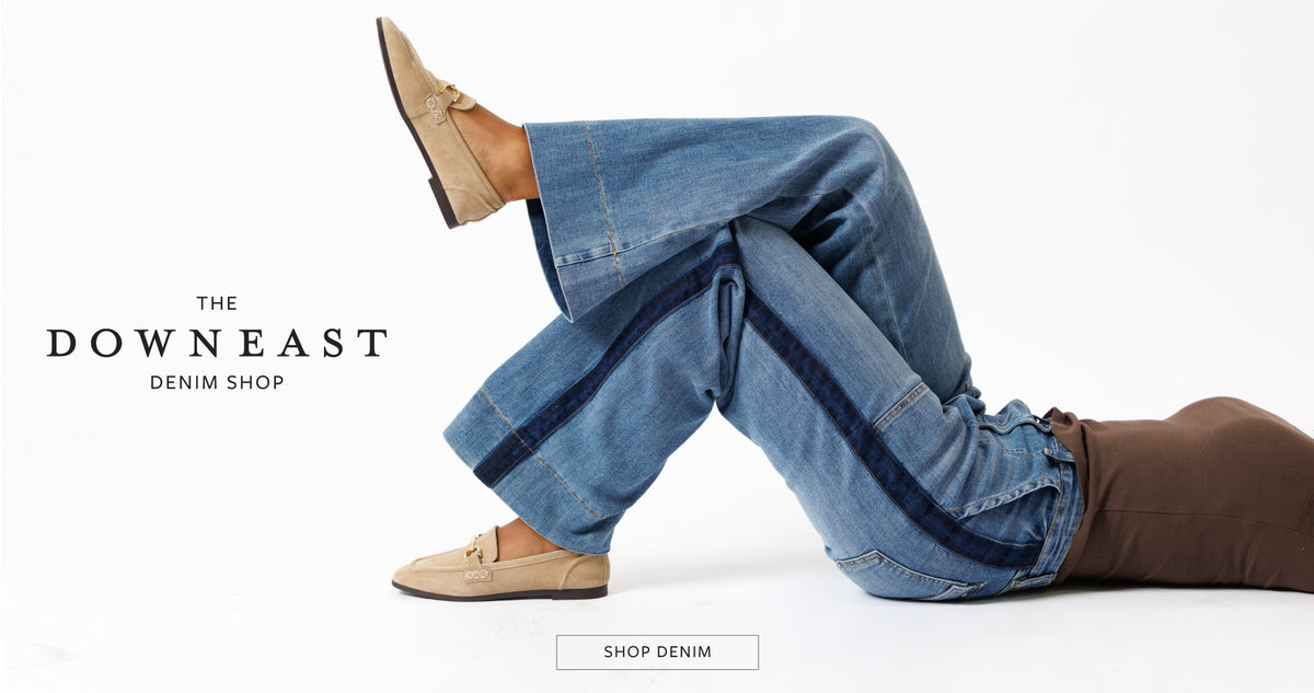 A person reclining on a white studio floor wearing loose blue denim jeans with dark contrast stitching and tan loafers. The text 'THE DOWNEAST DENIM SHOP' appears on the left, with a 'SHOP DENIM' button at the bottom.