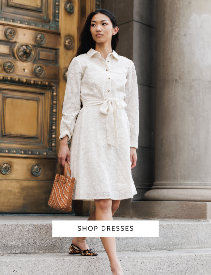 A woman stands on stone steps in front of a large bronze door and stone column, wearing a light cream long-sleeve collared dress with a tie waist and carrying a brown patterned tote bag. A white banner at the bottom reads 'SHOP DRESSES'.