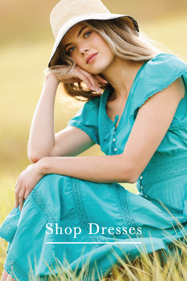 A woman wearing a turquoise blue dress and a beige wide-brim hat sits in a grassy field. The text 'Shop Dresses' is overlaid at the bottom.