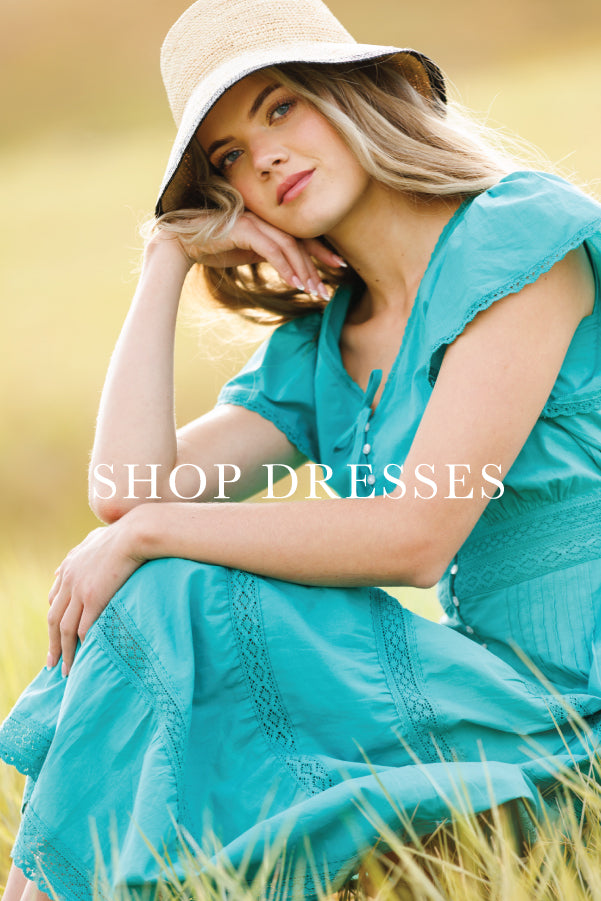 A woman wearing a turquoise short-sleeve dress and a beige wide-brim hat sits in a grassy field. A semi-transparent watermark reading 'SHOP DRESSES' is overlaid across the center.
