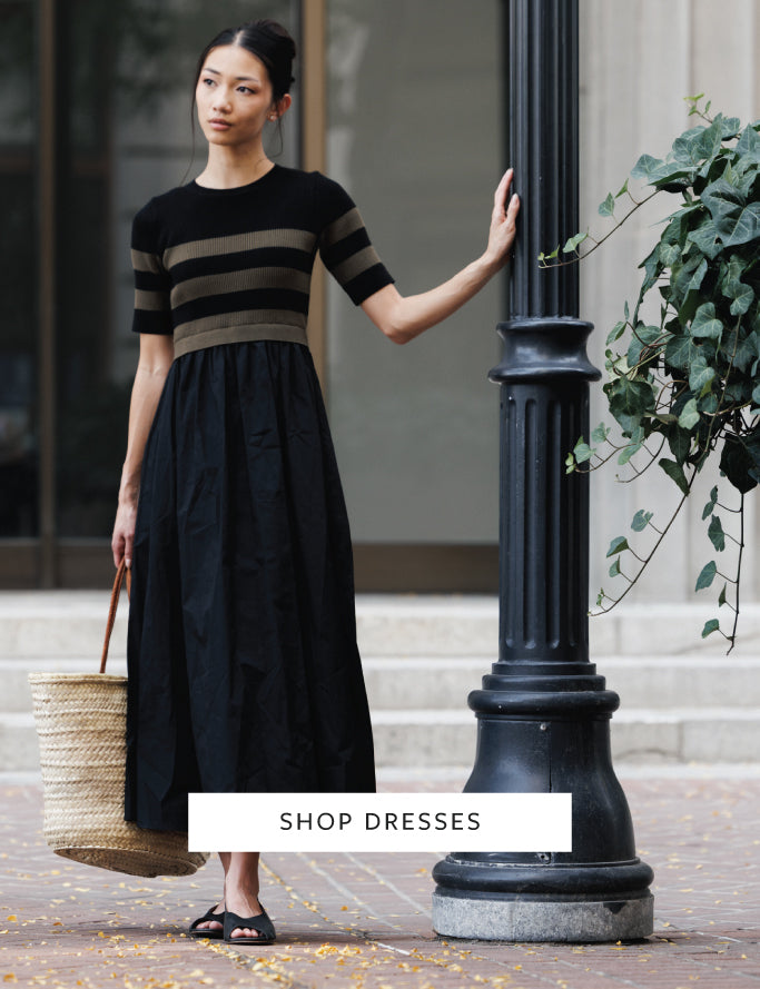 A woman stands outdoors beside a black lamppost wearing a black midi dress with beige horizontal stripes across the chest and sleeves. She holds a woven straw tote bag.