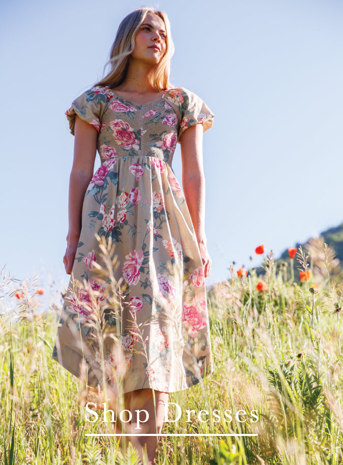 A woman wearing a beige floral midi dress stands in a sunlit grassy field under a clear blue sky. The dress has short flutter sleeves, a scoop neckline, and a pink and red floral print.