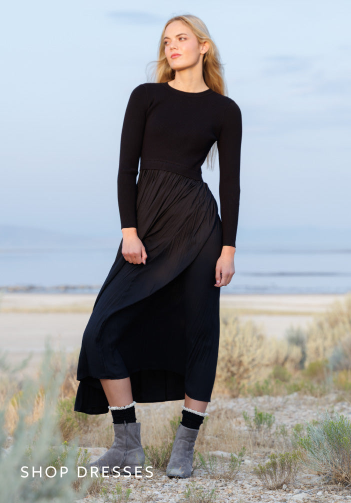 A woman stands outdoors in a coastal or desert-like setting wearing a long-sleeve black midi dress with a pleated skirt and gray ankle boots. The text 'SHOP DRESSES' appears in the lower left corner.